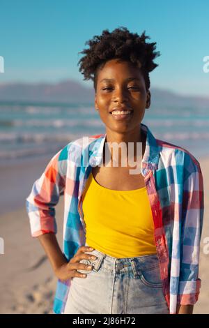 Porträt einer glücklichen afroamerikanischen jungen Frau mit kurzen Haaren und Hand auf der Hüfte am Strand gegen den Himmel Stockfoto