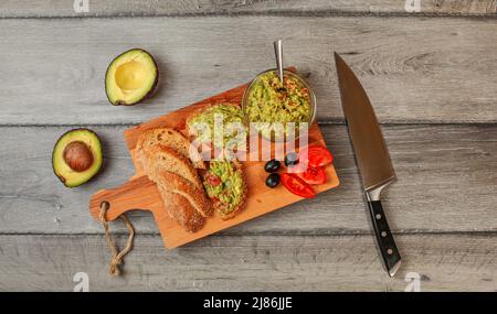 Flaches Lay Photo, frisch zubereitete Guacamole in kleiner Glasschüssel, Brot, Tomaten, Oliven am Arbeitstisch und zwei Avocados am nächsten grauen Holzschreibtisch Stockfoto