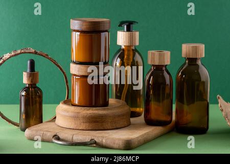 Braune Glasbehälter für Kosmetika mit Holzdeckeln. Cremedose, Tropfflasche und Lotion. Grüner Hintergrund. Pflege und Schönheit. Flaschen. Umweltfreundliches Konzept. Kosmetische Set-Box. Pipette und Spender. Stockfoto