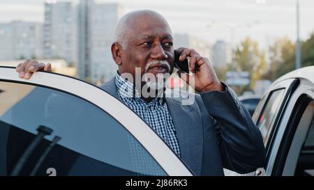 Reifer afroamerikanischer Geschäftsmann, der im Freien auf dem Parkplatz in der Nähe des Autos steht, spricht auf dem Mobiltelefon älterer, freundlicher Mann kommuniziert am Telefon Stockfoto