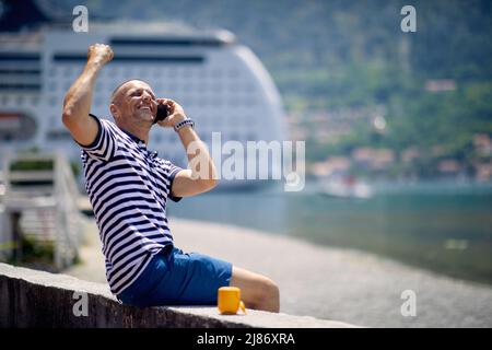 Mann fühlt sich siegreich, jubelt beim Telefonieren, sitzt auf Deck. Natur, Kreuzer und Berge Hintergrund. Reise-, Business- und Lifestyle-Conce Stockfoto