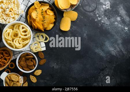 Ungesunde Lebensmittel. Snacks. Alle klassischen Kartoffelsnacks mit Erdnüssen, Popcorn- und Zwiebelringen und gesalzenen Brezeln in Schüsseln auf schwarzem Beton-Backgroun Stockfoto