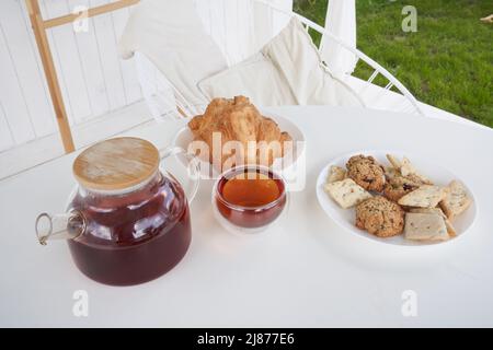 Sonntagsfrühstück im Hinterhof der Hütte. Frische süße Croissants mit Keksen auf dem Tisch. Transparente Teekane aus Glas mit gebrühtem Tee. Hochwertige Fotos Stockfoto