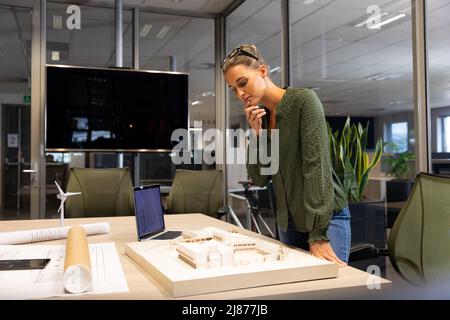 Kaukasische Architektin untersucht Architekturmodell auf dem Schreibtisch am modernen Arbeitsplatz Stockfoto