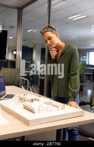 Nachdenkliche kaukasische Architektin untersucht Architekturmodell auf dem Schreibtisch am modernen Arbeitsplatz Stockfoto