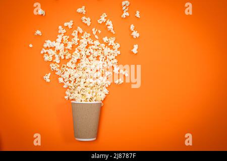 Popcorn wird aus dem Glas auf einem orangefarbenen Hintergrund ausgestreut, von oben betrachtet. Ein Spritzer Popcorn Stockfoto