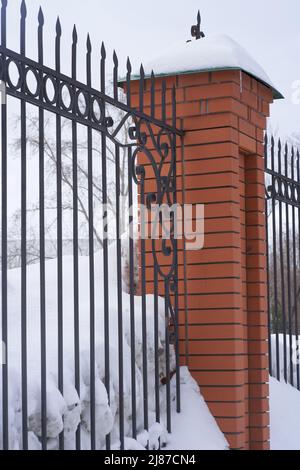 Nahaufnahme eines Metallzauns mit Ziegelsteinsäulen. Vor dem Hintergrund von weißem Schnee an einem Wintertag. Hochwertige Fotos Stockfoto