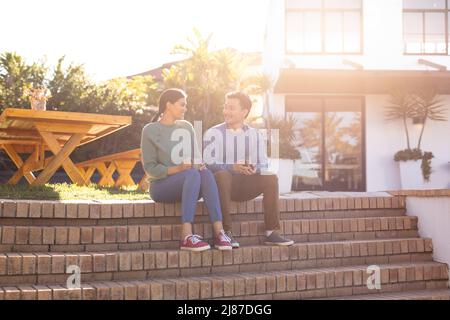 Liebevolles junges asiatisches Paar, das Kaffee hat, während es vor dem Haus auf einer Treppe sitzt Stockfoto