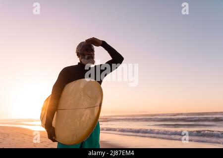 Bärtiger afroamerikanischer Senior mit Surfbrett, das die Augen am Strand vor dem klaren Himmel abschirmt Stockfoto