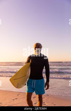 Bärtiger afroamerikanischer Senior mit Surfbrett, das bei Sonnenuntergang gegen Meer und klaren Himmel steht Stockfoto