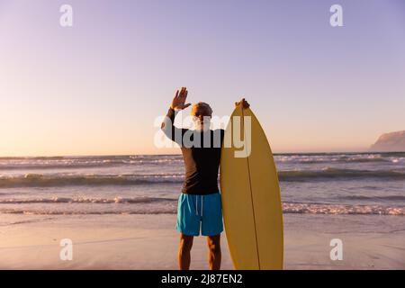 Bärtiger afroamerikanischer Senior mit Surfbrett, der vor dem Meer und dem klaren Himmel winkt Stockfoto