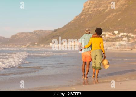 Rückansicht eines afroamerikanischen Paares mit den Armen, das bei Sonnenuntergang am Ufer gegen den klaren Himmel läuft Stockfoto