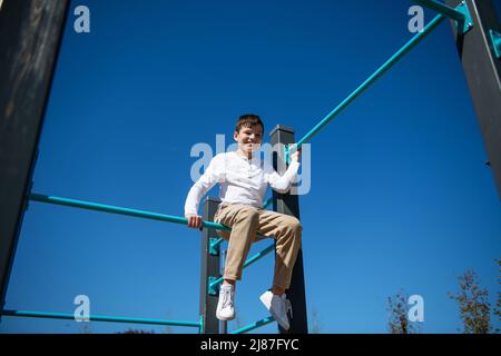 Ein Teenager-Junge sitzt auf einer horizontalen Stange. Ansicht von unten. Himmel im Hintergrund. Stockfoto