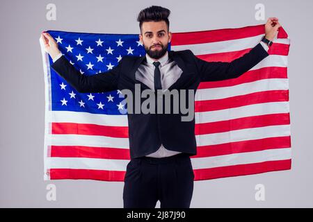 arabischer junger, schöner Mann mit schwarzem Haar im stilvollen Business-Anzug, der die amerikanische Flagge im Studio hält, isoliert weißen Hintergrund Stockfoto