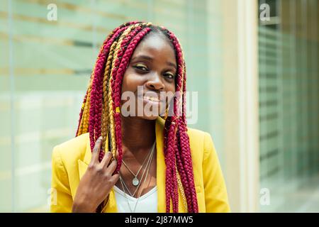 Kreative Stylistin afroamerikanische junge Frau mit Kanekalon Haar lange Afro-Zöpfe in einer gelben Jacke. Schöne Kleidung Designer Schneiderin posiert in Stockfoto