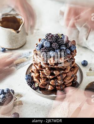 Stapel von Schokoladenwaffeln mit Heidelbeeren und Brombeeren. Das Konzept der Bewegung im Moment mit den Händen. Vertikale Ausrichtung Stockfoto