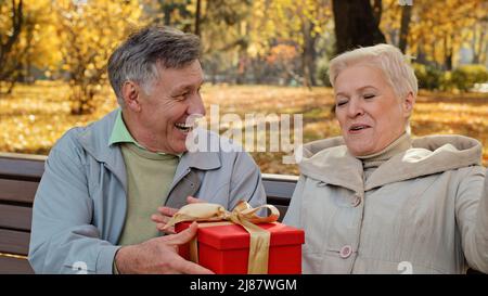 Reifer Mann gibt Geschenk an geliebte Frau am Geburtstag ältere Frau glücklich lacht positiv verheiratetes Paar feiert Jahrestag unerwartete Überraschung Stockfoto