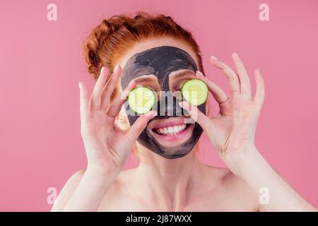 Rothaarige Ginger-Hündin mit Gurke und Tonmaske auf ihrem hübschen Gesicht, schneeweißes Lächeln auf rosafarbenem Hintergrund im Studio Stockfoto