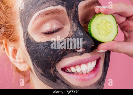 Rothaarige Ginger-Hündin mit Gurke und Tonmaske auf ihrem hübschen Gesicht, schneeweißes Lächeln auf rosafarbenem Hintergrund im Studio Stockfoto