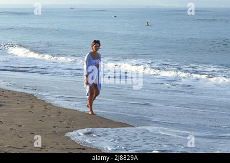 Eine junge weißgekleidete kaukasische Brünette, die am Strand von Batu Bolong Beach in Canggu, Bali, Indonesien, am Wasserrand läuft. Stockfoto