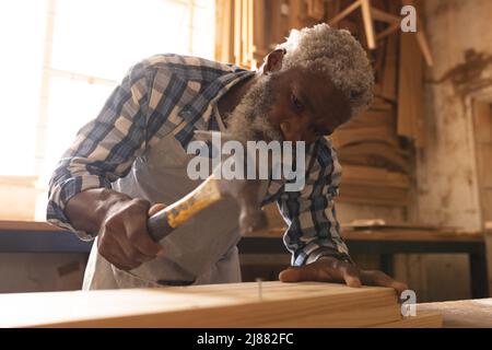 afroamerikanischer reifer Zimmermann, der in der Werkstatt Hammer auf Nagel in Plank einschlägt Stockfoto