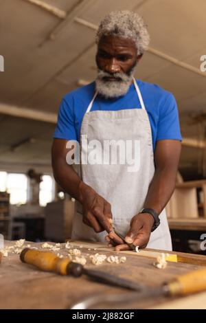 afroamerikanischer reifer Zimmermann, der Meißel auf der Planke auf der Werkbank in der Werkstatt verwendet Stockfoto