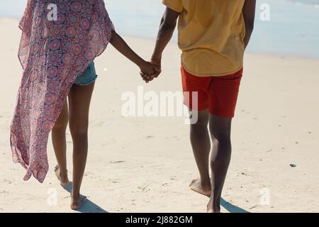 Rückansicht niedriger Abschnitt eines jungen afroamerikanischen Paares, das sich die Hände hielt, während es am Strand auf Sand ging Stockfoto