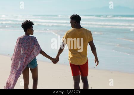 Rückansicht eines jungen afroamerikanischen Paares, das die Hände hielt und gegen den Himmel auf das Meer ging Stockfoto