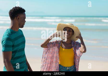 Lächelnde afroamerikanische junge Frau mit Hut und Sarong-Jacke, die ihren Freund am Strand ansieht Stockfoto