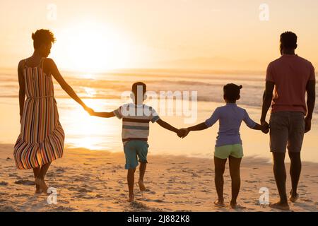 Rückansicht der afroamerikanischen jungen Eltern, die beim Strandspaziergängen die Hände von Tochter und Sohn halten Stockfoto