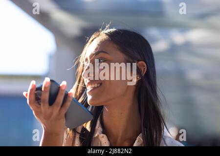Glückliche junge biracial Geschäftsfrau im Gespräch über Smartphone-Lautsprecher auf dem Flur an sonnigen Tag Stockfoto