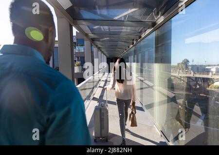 afroamerikanischer Geschäftsmann, der mit Gepäck auf dem Flughafenflur hinter einer Geschäftsfrau herläuft Stockfoto