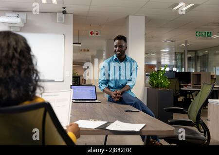 Glücklicher afroamerikanischer Geschäftsmann, der am Schreibtisch sitzt und mit einer Geschäftsfrau aus der Birazialwelt diskutiert Stockfoto