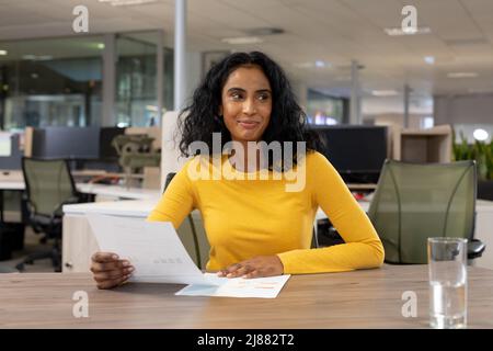 Lächelnde Geschäftsfrau mit Birazial, die mit einem Dokument am Schreibtisch sitzt, während sie auf den modernen Arbeitsplatz blickt Stockfoto