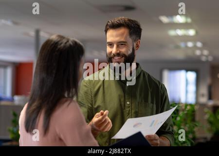 Lächelnder kaukasischer Geschäftsmann, der am Arbeitsplatz über ein Dokument mit einer biracial Geschäftsfrau diskutiert Stockfoto