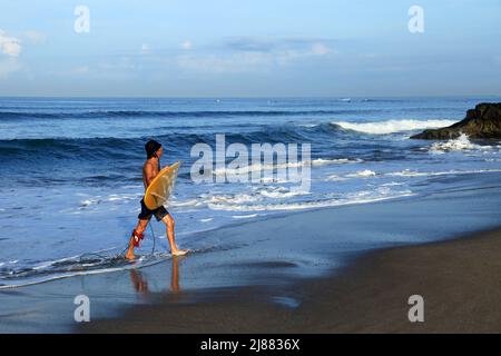 Ein kaukasischer Mann, der ein gelbes, langes Surfbrett trägt und das Meer mit Wellen im Hintergrund am Batu Bolong Beach, Canggu, Bali, verlässt. Stockfoto