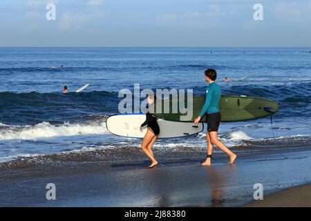 Ein kaukasisches Paar, das mit Longboard-Surfbrettern am Batu Bolong Beach in Canggu, Bali Indonesien, ins Meer geht, mit Surfern im Hintergrund. Stockfoto