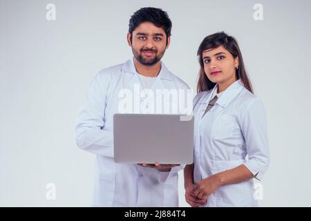 Schöne indische Frau und hübscher putenmann in weißem Blazer-Mantel mit Laptop und Blick auf die Kamera im Studio-Hintergrund Stockfoto