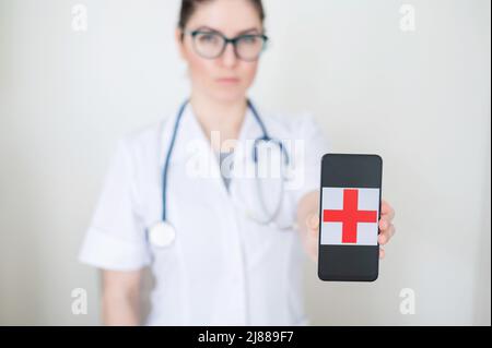 Ärztin in Brille hält ein Smartphone mit einem roten Kreuz auf dem Bildschirm. Ernsthafte Krankenschwester in einem medizinischen Mantel mit einem Stethoskop auf seinen Schultern Stockfoto
