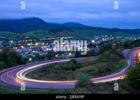 Wolkiger Abend auf einer Bergstraße in den ukrainischen Karpaten. Spuren von Scheinwerfern von sich schnell bewegenden Autos. Kleines Dorf mit Lichtern im val Stockfoto