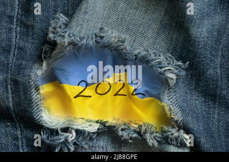 Die Flagge der Ukraine und der Text 2022 sind auf einer Papierkarte aus einem Loch in Jeans, Mode und Trend, Krieg in der Ukraine im Jahr 2022 gesehen gezogen Stockfoto