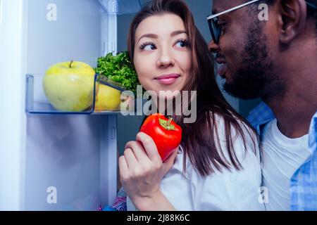 Blick auf den Kühlschrank:afroamerikaner Mann mit asiatischer Frau nehmen ein Gemüse mit offener Tür Stockfoto