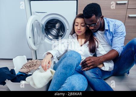 afroamerikanischer Mann hilft glücklich asiatische Hausfrau zu verwenden Waschmaschine drinnen, Umarmung und Zärtlichkeit Stockfoto