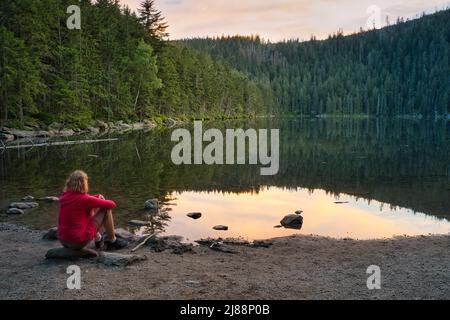 Eine kaukasische Frau mit blonden Haaren und einer roten Jacke, die am Ufer des Sees Certovo jezero im Böhmerwald, Tschechische Republik, sitzt. Nach Sonnenuntergang. Stockfoto