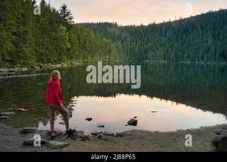 Eine kaukasische Frau mit blondem Haar und einer roten Jacke, die am Ufer des Sees Certovo jezero im Böhmerwald, Tschechische Republik, steht. Nach Sonnenuntergang. Stockfoto