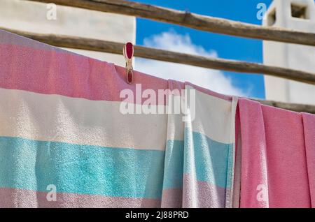 Bunte Badetücher, die mit einer Wäscheklammer an einer Wäscheleine hängen. Selektiver Fokus. Wolkiger Himmel und Kamin im Hintergrund. Stockfoto