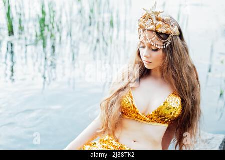 Fantasy Frau echte Meerjungfrau mit Dreizack Mythos Göttin des Meeres mit goldenen Schwanz sitzt in Sonnenuntergang auf Felsen.. Gold Haar Krone Muscheln Perlen Schmuck. Merma Stockfoto