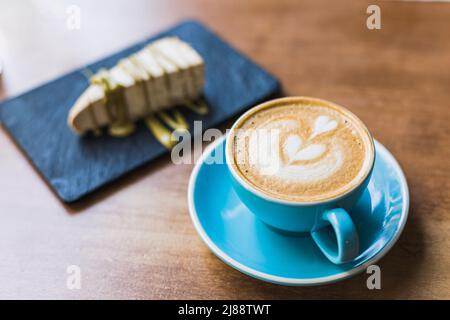 Köstlicher Kaffee mit Latte Art Herz in einer blauen Tasse auf einem Holztisch geformt. Im Hintergrund ein Stück Käsekuchen mit Pistazie Stockfoto