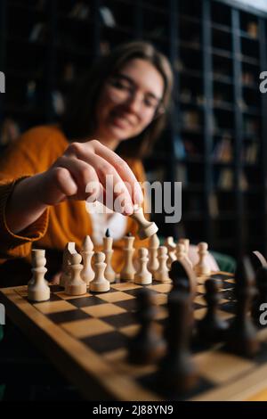 Vertikale, selektive Fokusaufnahme einer lächelnden Frau mit eleganten Brillen, die Schach in Bewegung bringen und auf einem Sessel im dunklen Bibliotheksraum sitzen lässt. Stockfoto