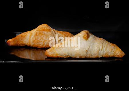 Zwei gebackene Croissants auf Tablett im elektrischen Ofen zu Hause, schwarzer Hintergrund Stockfoto